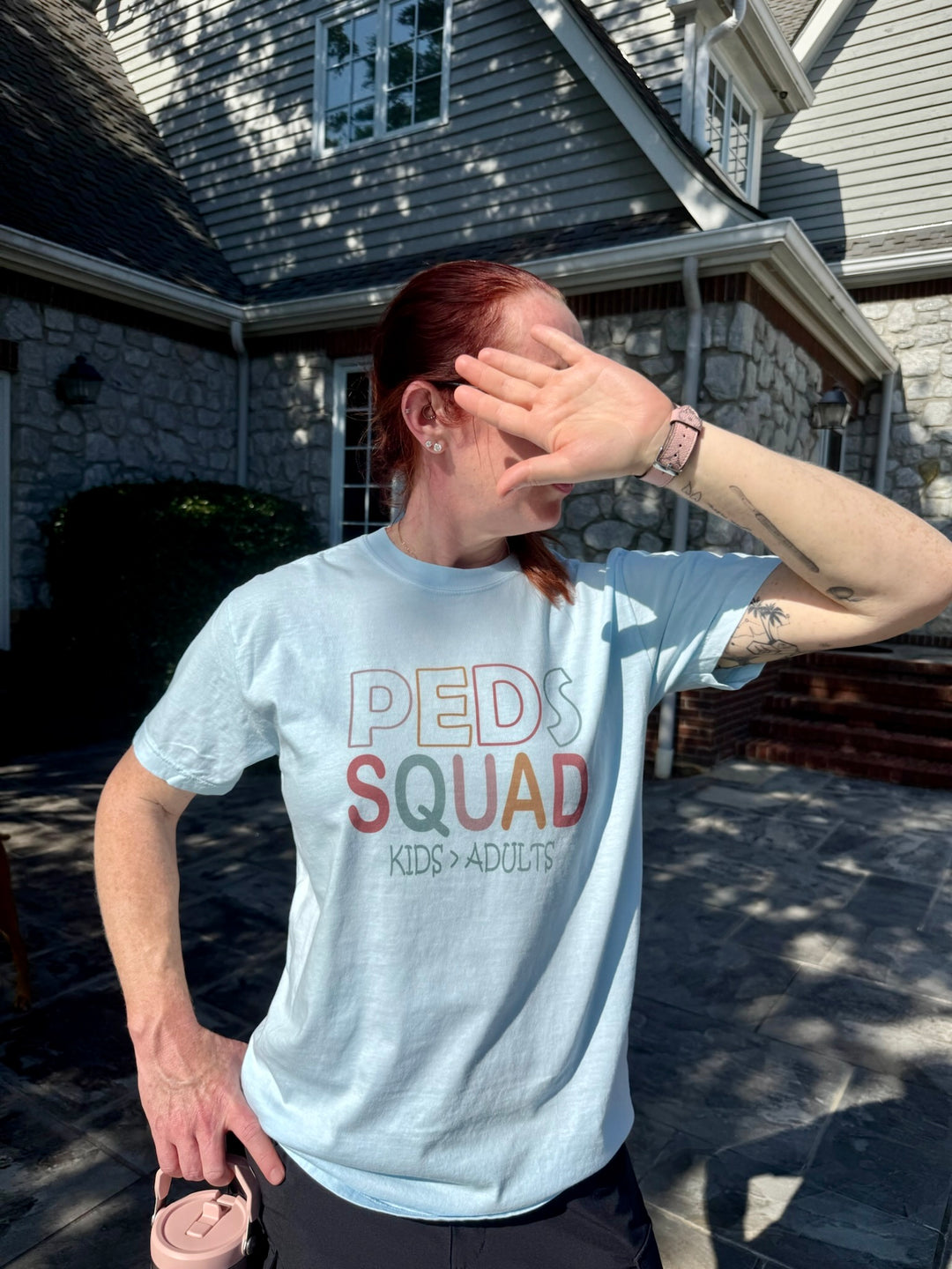Dr. Betsy Grunch raises a hand to shield her eyes from the sun while wearing the “PEDS SQUAD – KIDS > ADULTS” tee. She stands on a stone patio in front of a gray house with stone siding.