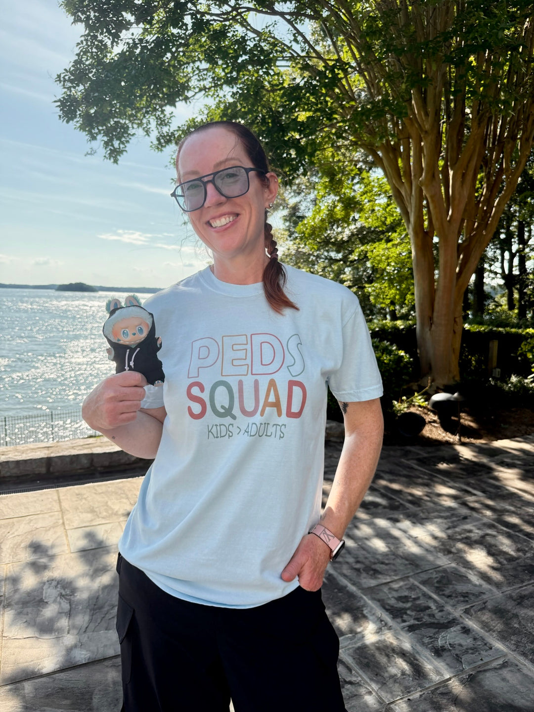 Dr. Betsy Grunch smiles and holds a small doll while modeling the “PEDS SQUAD – KIDS > ADULTS” shirt on a patio near the lake, surrounded by trees and water.